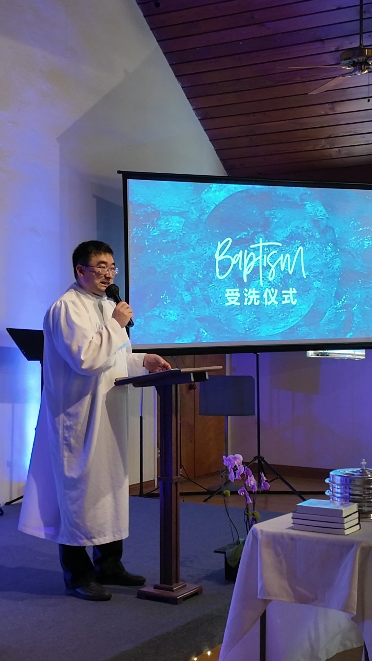 牧师在讲台前主持受洗仪式，屏幕上写着受洗仪式