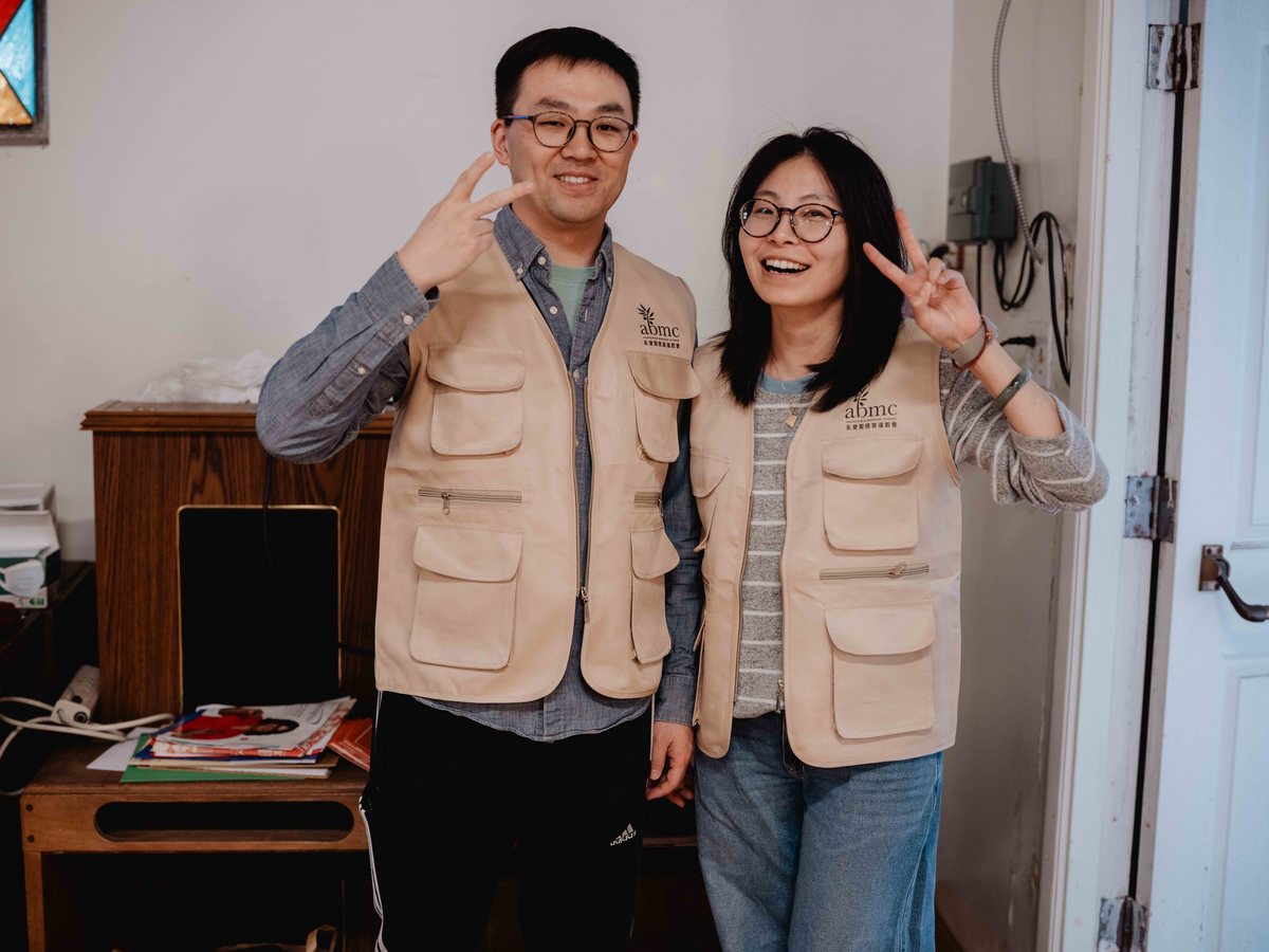 两位穿着教会背心的同工在门口比出手势，笑着迎接