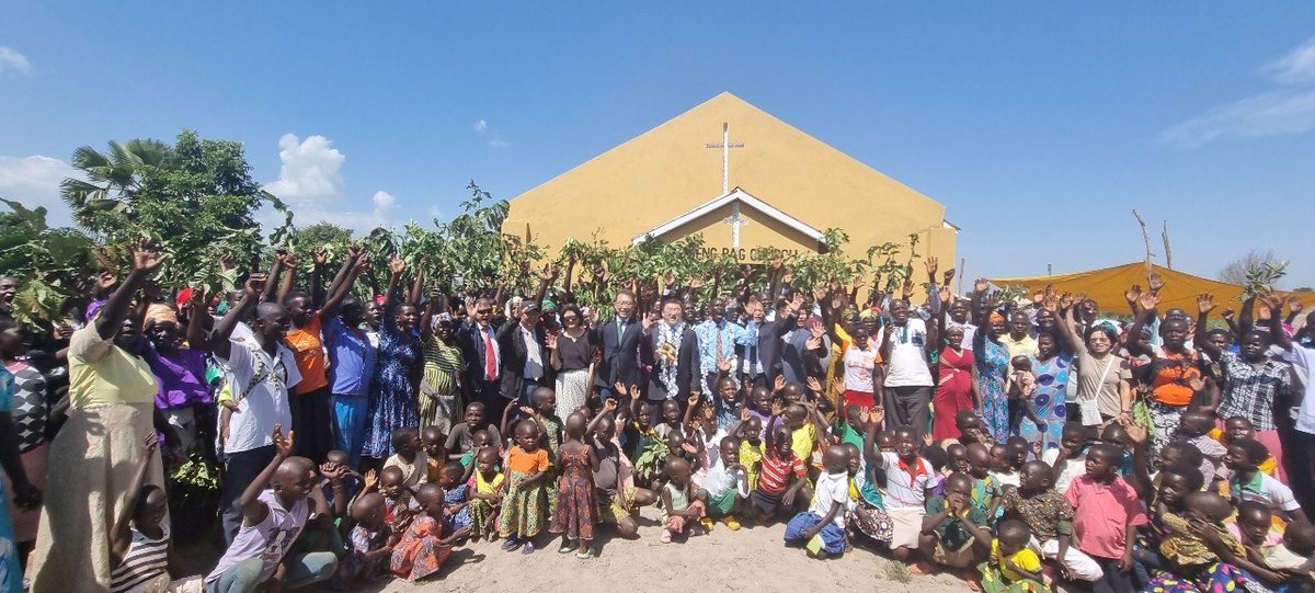 A large group photograph outside a village church, adults and children with their hands in the air